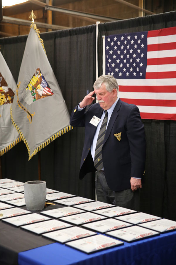 A man standing in front of an American flag saluting toward a table.