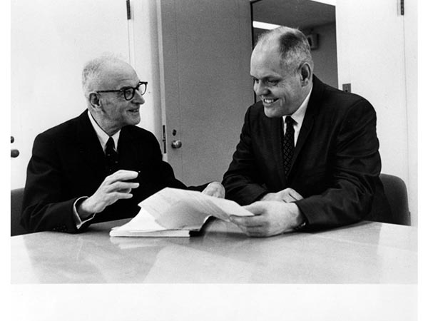 A black and white photograph of two men seated at a table looking at a document.