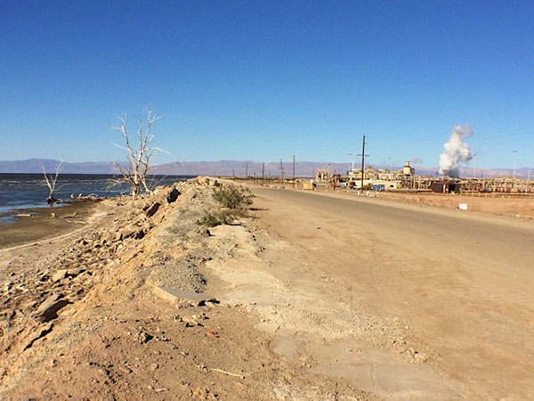 At the shore of the Salton Sea, a road divides two very different views. On one side, blue water and bleached trees captured by countless nature photographers; on the other, one of many geothermal plants that promise clean energy and local jobs.