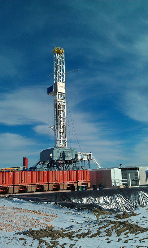 A tall white drill tower stands on and orange rig, with mountains and a partly cloudy blue sky in the background.