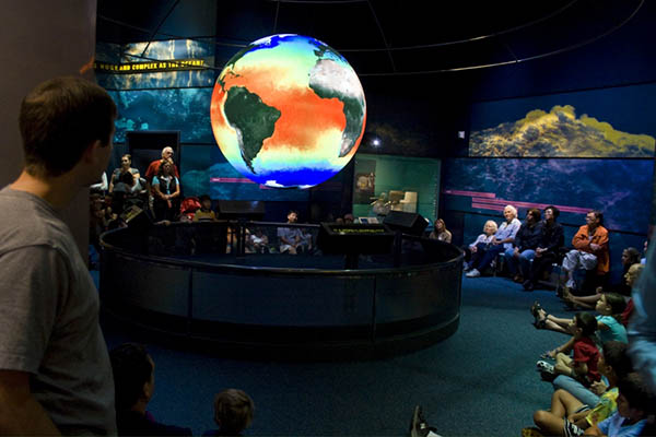 Photograph of a crowd sitting and standing around and watching a model of the earth. 