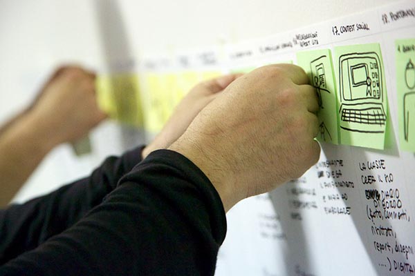 Photograph of hands putting sticky notes with drawings on them onto a grid hung no a wall.
