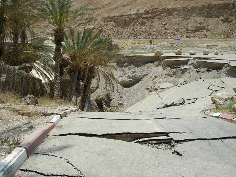 Photo of a cracked paved road, ending in a large sinkhole, with palm trees falling in.