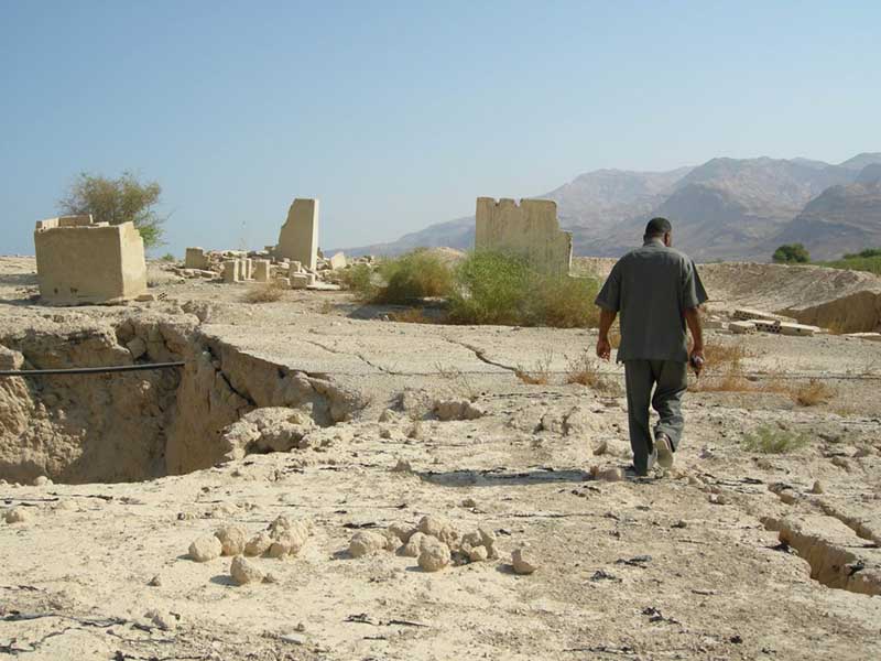 A figure walking across a dusty landscape, with a large sinkhole on the left, and fallen buildings in the distance, mountains beyond.