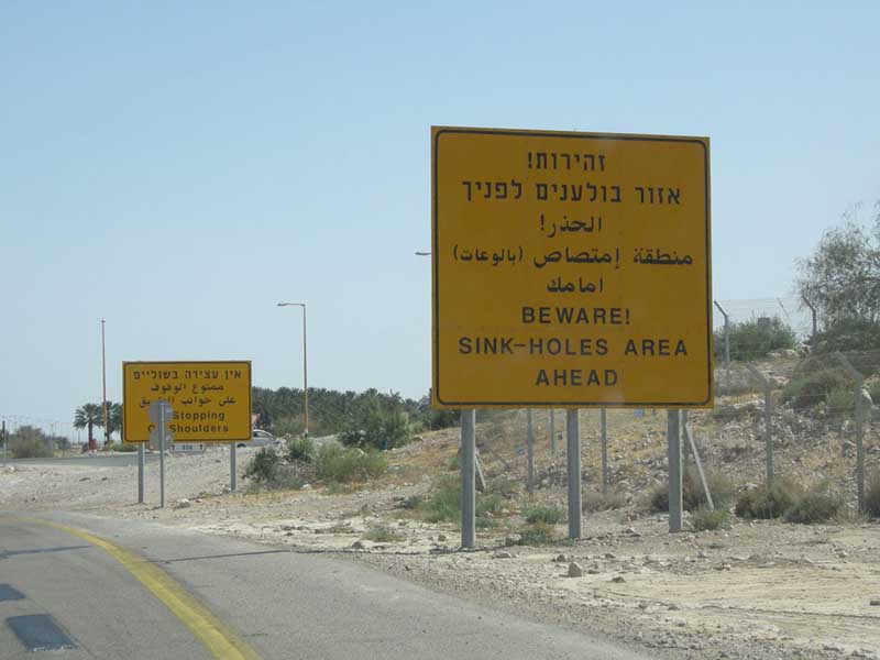 Photo of two signs at a roadside, reading "Beware: Sink-Holes Area Ahead," in Hebrew, Arabic, and English.