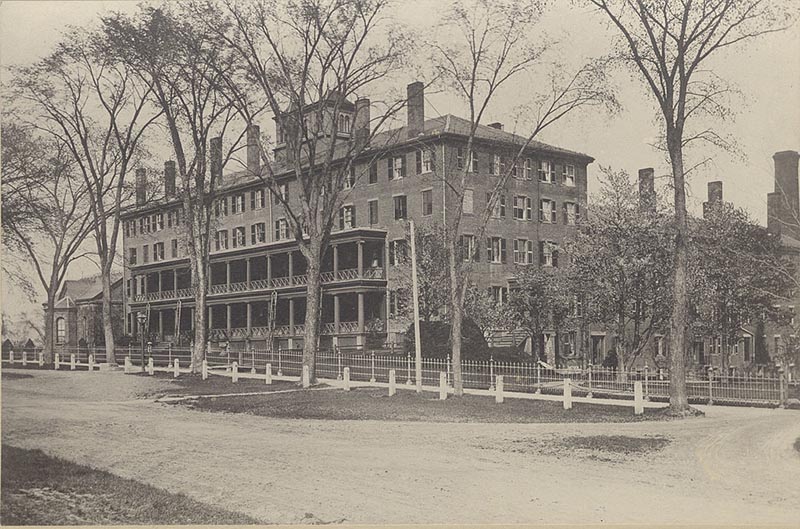 Faded photograph of an historic rectangular brick building, with trees, white fence, and a dirt road in front.