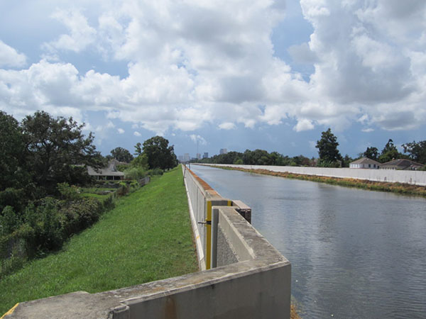 Photo of a canal on the right, with concrete sides, a green embankment on the left, and a partly cloudy sky.