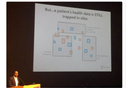 Photograph of a presentation slide projected on a screen, with the speaker in the lower left hand corner at a podium. Slide says: "But...a patient's health data is STILL trapped in silos," and shows grey squares with smaller blue and orange squares that appear to represent siloed data.