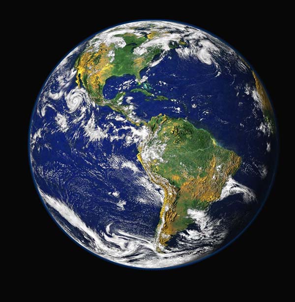 Classic image of the earth from space, like a large blue marble against a black background. South America is slightly to the right and North America up and to the left. White clouds swirl over blue oceans and green and yellow/orange land.