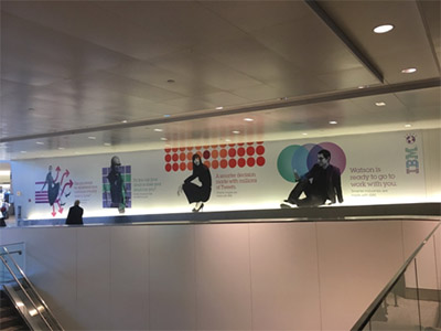 Billboard ad on the side wall of an airport, white background with silhouettes of people sitting; IBM logo on right; graphical images such as a grid of red dots behind one figure of a person squatting on heels.