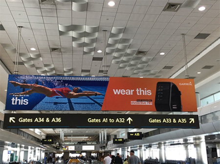 Billboard hanging from an airport ceiling over a sign for different gates, with what looks like a moving walkeway below, windows on left and right, and the tops of travelers' heads. On left, billboard shows picture of a swimmer diving into a lap pool; left says "wear this" and shows a black device or watch on an orange-red background.