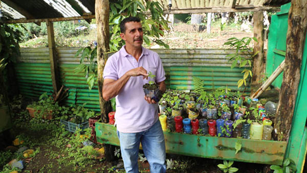 A man in a lavendar short-sleeved shirt and jeans stands in a gardening shed full of plants in small containers, and what look like colored upside-down plastic bottles. He is holding a small planter with a green shoot sprouting a few leaves.