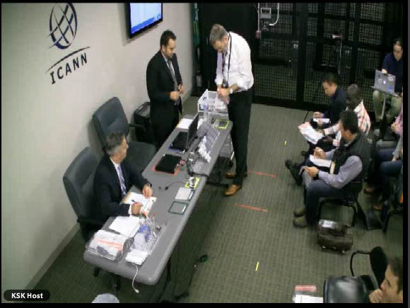 Screen shot still image of two men seated behind a grey table in the left half of the picture, with laptops and other papers and equipment. Behind them is a white whall with the ICANN name and logo. A small flatscreen can be seen as well. Across from them, on the right side, are people seated like an audience, possibly taking notes. About three or four are visible but possibly more sit behind them.