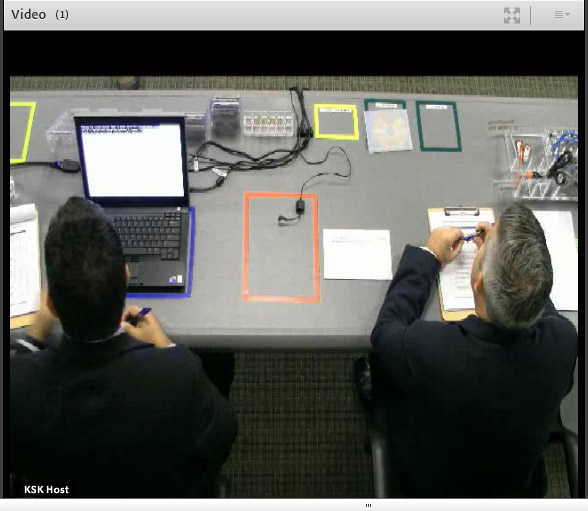 Image of two men, in what look like suit jackets, seated at a grey table (taken from above and behind them). On the table are various taped-off squares in orange, blue, yellow, etc., and miscellaneous paper. In one blue square is an open black laptop.