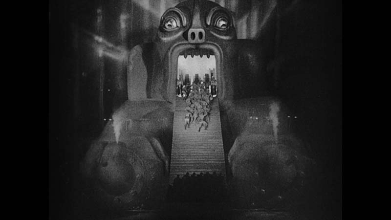 Black and white still from Metropolis, showing a large mechanical demon head with stairs for a tongue, and workers on the stairs.