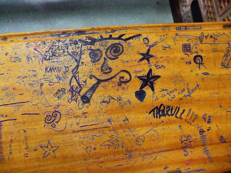 A wooden desk covered in graffiti drawn with blue ballpoint pen, including a face smoking a joint, a star, and writing in French. 