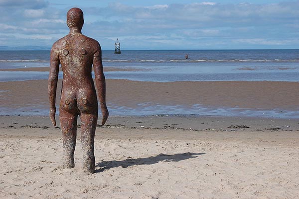 An iron sculpture of a human form, photographed from behind, stands on an ocean beach looking out to sea. Its legs are embedded in the sand, up to the knees. In the distance, just past the tide line, another figure stands in the water. There is a buoy further out in the water. 