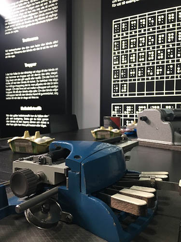 A bulky machine with a few keys sits on a large black table, with other supplies on it in the background, and a large informational poster about braille hangs on the wall behind it.