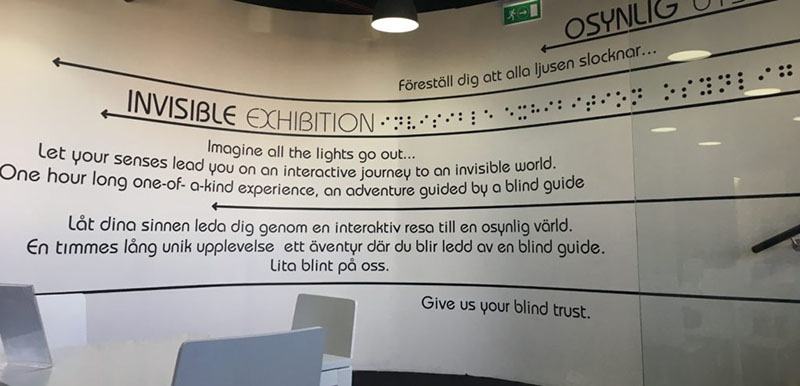The picture shows a white wall filled with text that is right as you come into the exhibition. It is an advertisement for the exhibition and prepares the visitor for their guided tour. It reads "Imagine the lights go out... Let your senses lead you on an interactive journey to an invisible world. One hour long one-of-a-kind experience, an adventure guided by a blind guide... give us your blind trust."