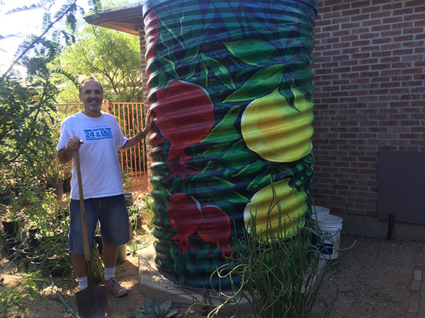 A man stands next to a large cistern painted with leaves and fruit. A brick building is behind the cistern.
