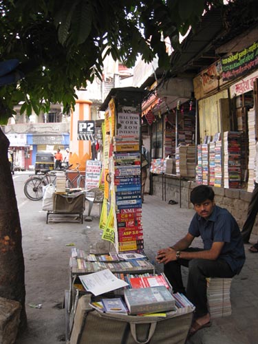 A man sits by piles of books for sale on a sidewalk.