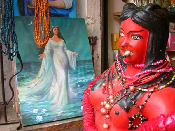 In the foreground is a sculpture of a red woman with black hair smoking a cigar. In the background is a painting of a woman in a long flowing dress.