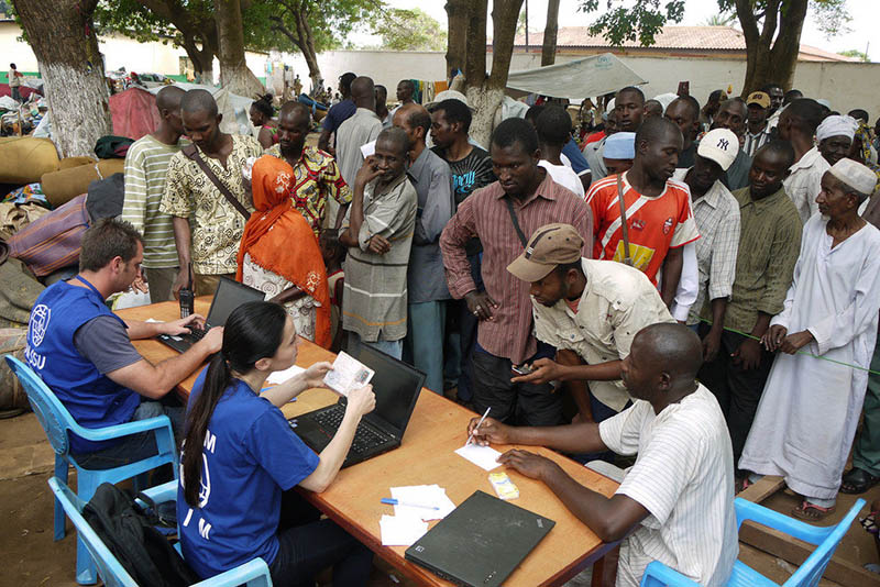 A white man and woman in blue t-shirts are seated at a table with laptops. A crowd of African people are standing in front of them. On the right, an African man is seated filling out paperwork.