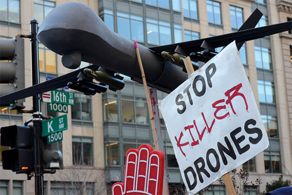 A black model drone plane is held up among protest signs that say "Stop killer drones."