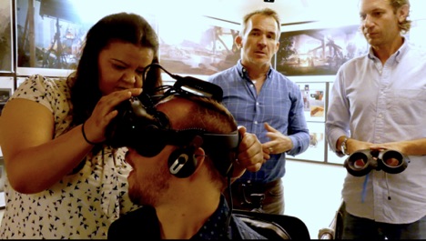 The image shows four people: Professor Tom Boellstorff is standing in the middle of the image but in the background. Two others are on the left and the right. The woman on the right is fitting a virtual reality headset to a man who has Parkinson's disease. He is seated and she stands. On the right hand side of the man in the chair, is another man. He is standing and hold the handsets that go with the virtual reality headset.