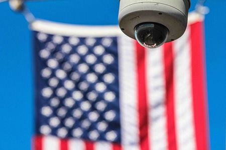 Photograph of a surveillance camera in focus. An American flag is hanging in the out of focus background.