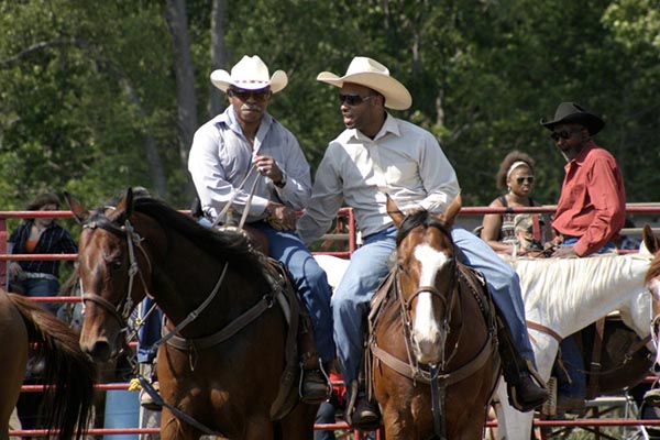 Photograph of men in Stetson style hats, white shirts and blue pants on horse back. More people and horses can be seen in the background.