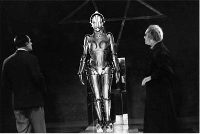 Black and white still. Two men in suits face a chrome female robot figure.