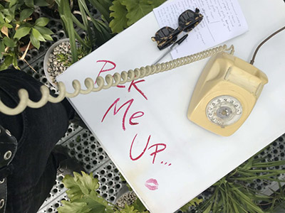 Photograph of a vintage yellow dial phone sitting on top of a white table that says "Pick me up..." on it with a lipstick kiss mark below it.