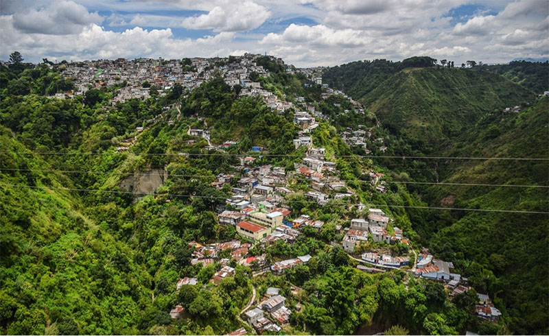 The picture shows a lush, green ravine/valley full of terra cotta roofed houses in Guatemala City's Zone 3