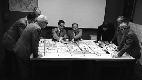 A group of white men meet around a table in a black and white photo. They are playing what looks to be a board game.