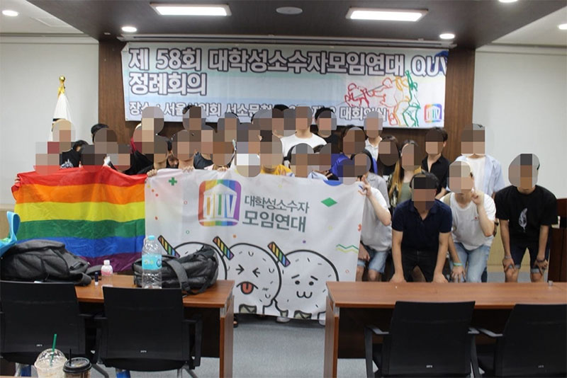 Photograph of a large group of people holding up banners and a rainbow flag. Their faces have been digitally obscured.