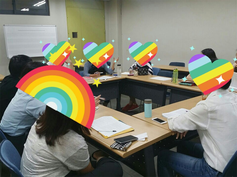 A group of South Korean activists sit around a square table. All of their faces are obscured by virtual "stickers," in the shapes of rainbows and rainbow hearts.