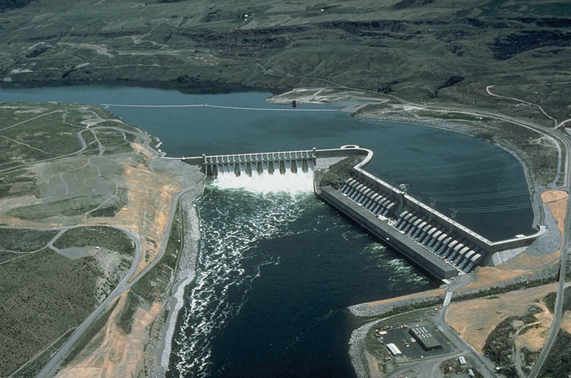 This image is an aerial view of a large hydropower dam. The upper half depicts the reservoir, the lower half shows the water gushing through the dam.
