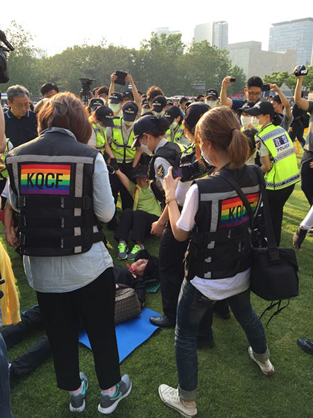 Picture of Queer Culture Festival Staff, police, protesters, and press documenting the police lift and carry anti-LGBT protesters from the ground.