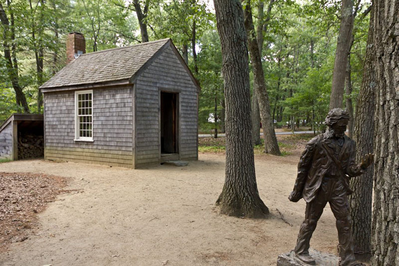  A small replica of Thoreau's cabin and statue near Walden Pond can be seen against a backdrop of green trees. The house is small, grey, one-room building with a red brick chimney. The statue is brownish in color, perhaps made of bronze.