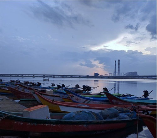 Red and white fishing boats sit along the Ennore coast. Behing them, we see a bridge in the distance and a stormy evening sky.