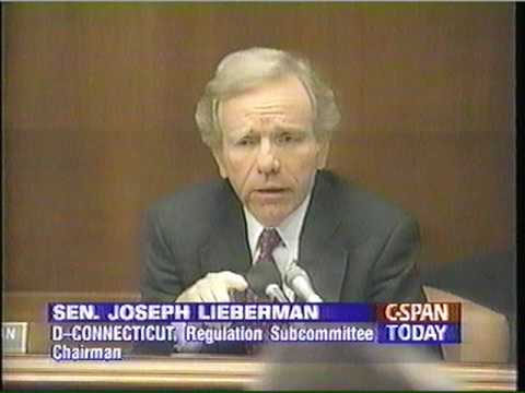 Still of Joseph Lieberman wearing a suit sitting at table talking into a microphone.