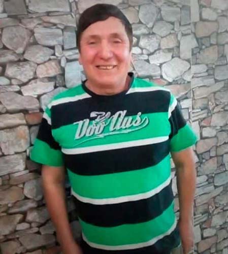 A stocky man in his 60's looks into the camera. He has short, brown hair swept to the right and olive skin. He is wearing a T-shirt with large green and black stripes interspersed with white stripes. The T-shirt reads "Doo Aus." Behind him is a stone wall filling the entire background of the photo.