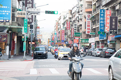 The image depicts taiwanese traffic, with a motorist in the foreground and cars in the background. Also in the background are buildings full of billboards, streetlights. The image conveys a general sense of both bustling street noise and crowded living/business quarters.