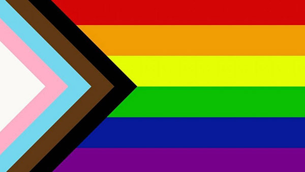 A rainbow flag (top to bottom red, orange, yellow, green, dark blue, and purple) is shown with 5 color chevron pattern coming from the left side, featuring black, brown, light blue, light pink, and white stripes.