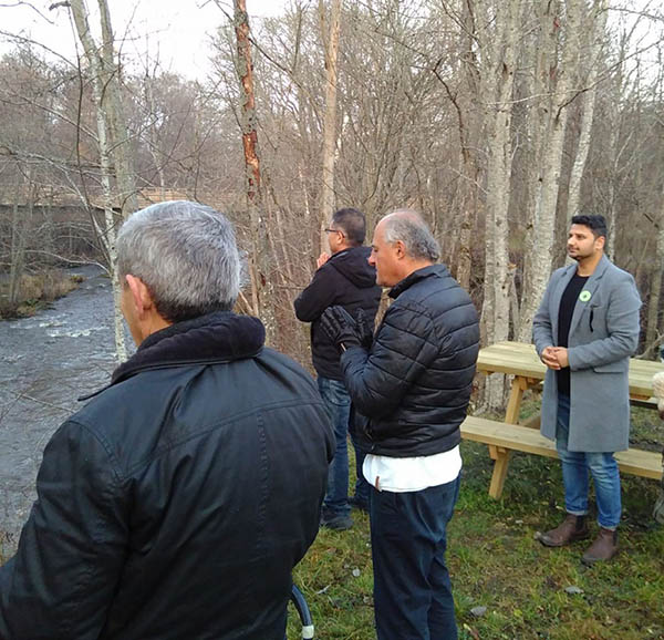NBI participants stand on near a stream and picnic table in the midst of some deciduous trees, eyes closed and hands clasped, listening.