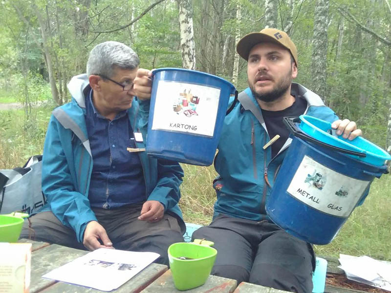 An NBI guide seated at a wooden table in the woods holds up two blue bins as example of recycling or waste containers.