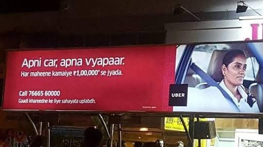 A photograph of a billboard. The left side is red with text. The right side is a photograph of a woman in a car. Between the two is the Uber logo.