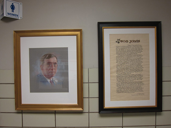 Photograph of a framed drawn portrait of a man. To the right of it is a framed page of calligraphy text titled Tom Jones.