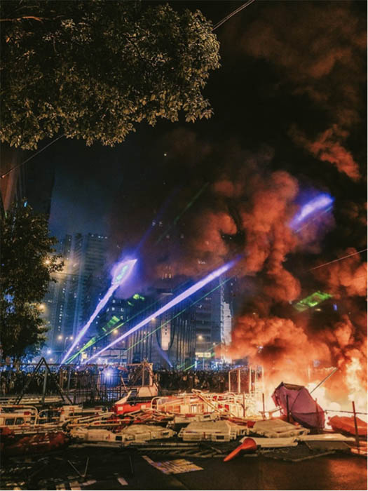 A city scene with fire and knocked over fencing in the foreground. Green and purple light beams emanate from a crowd in the background. Photo Credit: Deacon Lui, @lsb.co on Instagram